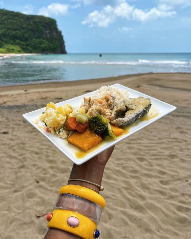 A plate of creole fish, rice, pumpkin and provisions held up on the beach